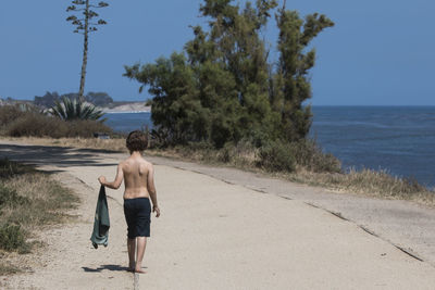 Rear view of shirtless man walking on road by sea