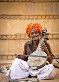 Portrait of man in traditional clothing playing sitar against wall
