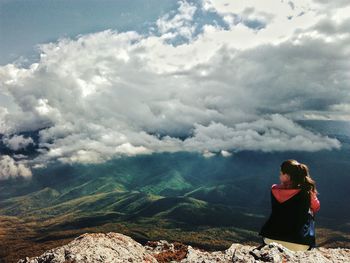 Rear view of young woman sitting on mountain against cloudy sky