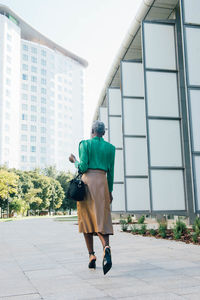 Full body back view of african american female in trendy outfit and high heels walking on paved walkway in city