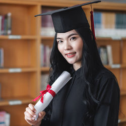 Portrait of student holding degree while standing against shelf