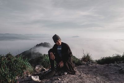 Young man sitting on rock against sky