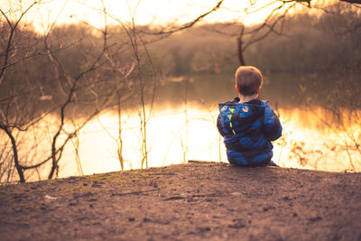 Rear view of boy sitting at lakeshore during sunset