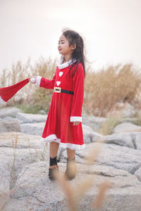 Girl wearing red dress holding santa hat while standing on rocks against sky