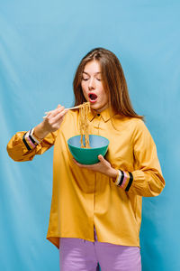 Positive young female in yellow shirt opening mouth to eat yummy instant noodles with chopsticks on blue background