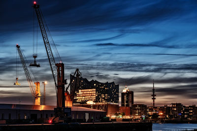 Cranes and buildings in city against cloudy sky at sunset