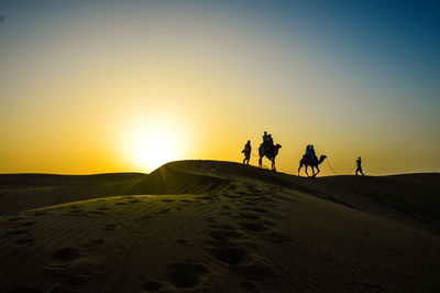 Silhouette people on camels at desert against clear sky during sunset