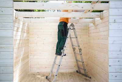 A male construction worker climbs a ladder to reach the roof of a wooden garden building. 