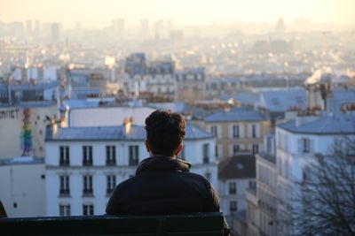 Rear view of man sitting on bench against cityscape