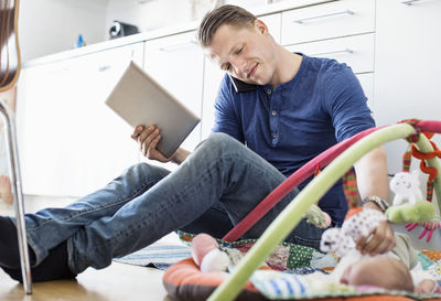 Father with digital tablet and mobile phone looking at baby lying on mat in kitchen