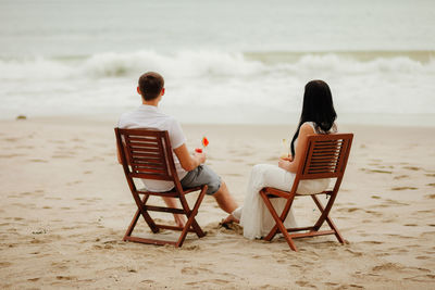 Rear view of couple having drink while sitting on chairs at beach