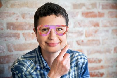 Portrait of smiling woman holding prop eyeglasses against wall