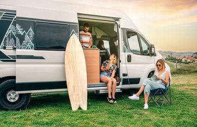 Two women sitting in front of camper van while young man cooks