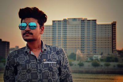 Portrait of young man wearing sunglasses standing against city