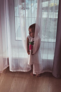 High angle view of cute toddler holding curtain while standing against window at home