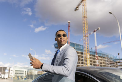 Businessman wearing sunglasses holding smart phone standing by car on sunny day