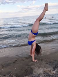 Young woman in bikini doing handstand at beach against cloudy sky