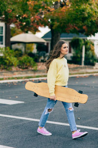 Girl in casual clothing crossing street holding a longboard as her hair blows in the wind