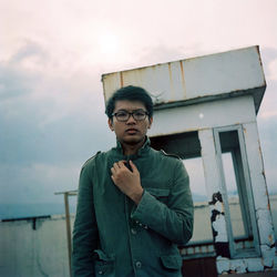 Portrait of young man wearing eyeglasses standing against cloudy sky