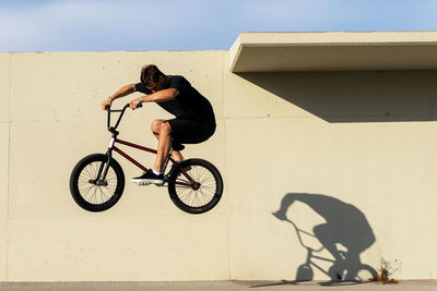 Full body side view of male in casual clothes jumping on bmx bicycle against sunlit concrete wall on summer weekend day on street