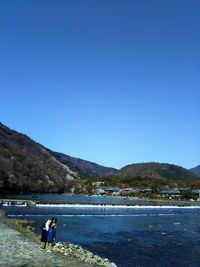 Rear view of friends standing by lake and mountains against clear blue sky