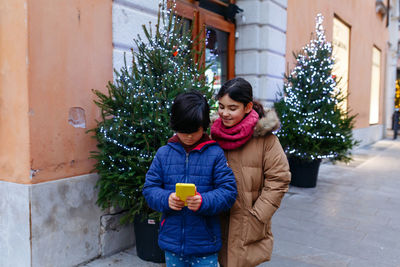 Two standing childreen on the street watching mobile phone together next to decorated christmas tree