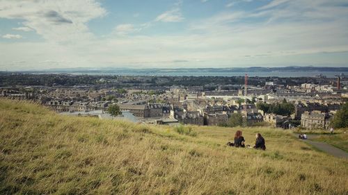 Women sitting on grass at calton hill over cityscape against sky