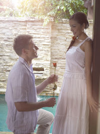 Man kneeling while proposing woman with red rose by swimming pool