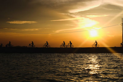 Silhouette people cycling at beach against sky during sunset