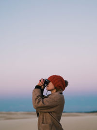 Side view of man looking through binoculars while standing against sky