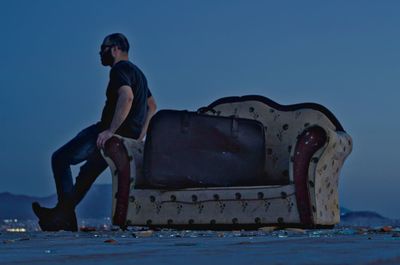 Full length of man leaning on abandoned sofa against clear sky at dusk