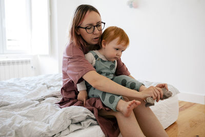 Mother puts sock to male toddler's foot sitting in bedroom