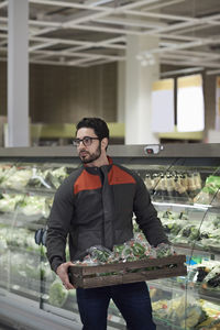 Sales clerk holding crate with green bell peppers by display cabinet while looking away at supermarket