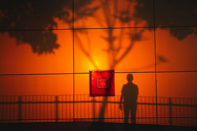 Rear view of silhouette man standing by railing against orange sky