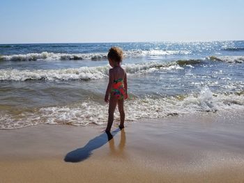 Rear view of girl in swimwear standing at beach against clear sky during sunny day