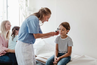 Female doctor examining boy's ear with otoscope while family sitting in medical room