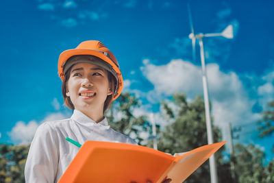 Female engineer in hardhat holding file while standing against windmill