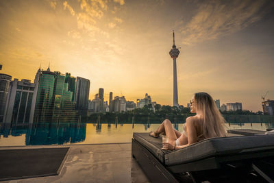 Woman sitting by buildings against sky during sunset
