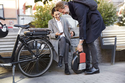 Smiling mature woman sitting on bench looking at man holding electric unicycle in city