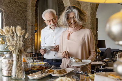 Smiling woman taking croissant from buffet standing by man at boutique hotel
