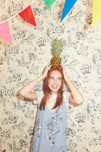 Smiling young woman carrying pineapple on head against wallpaper during party at home
