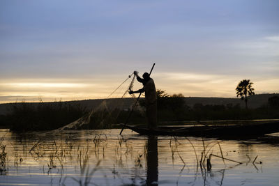Silhouette man fishing by lake against sky during sunset