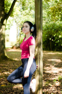Portrait of young woman listening music while standing against trees at park