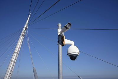 Low angle view of security camera by metallic cable against blue sky