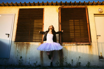 A ballerina in a tutu and a jacket stands against the wall of an old house pretending to take off