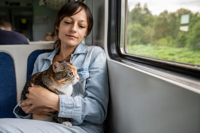 Public train ride of thoughtful smiling woman with calm peaceful tricolor cat sitting on lap.