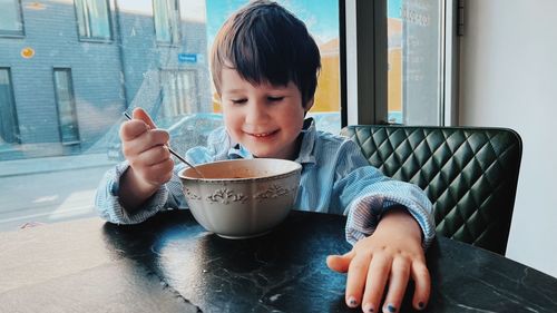 Smiling cute boy eating soup from a bowl while sitting at the big window in cafe