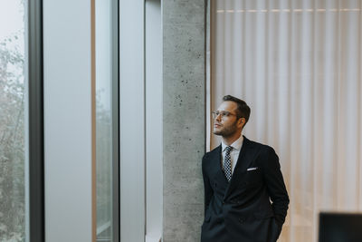 Thoughtful young businessman leaning on column while looking out through window at office