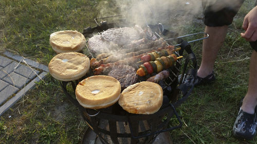 Low section of man cooking meat with vegetables and buns on barbecue grill in yard