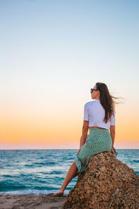 Rear view of woman sitting at beach against clear sky during sunset